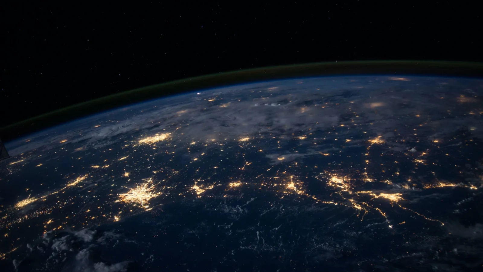 Vue de la Terre depuis l'espace, la nuit, montrant des villes et des régions illuminées, reliées par des lignes lumineuses, avec un ciel sombre et une faible lueur atmosphérique verte le long de l'horizon.