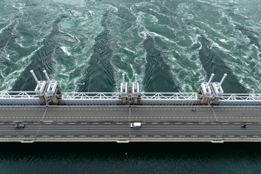 Vue aérienne d'un pont enjambant des eaux tourbillonnantes et turbulentes, avec quelques voitures traversant le pont et des courants visibles dans l'eau en contrebas.