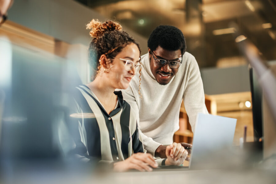 Two people collaborate at a desk, looking at a laptop screen. One is seated and typing, while the other stands beside them, leaning in and smiling as they discuss Instruments Support together in a modern office setting.