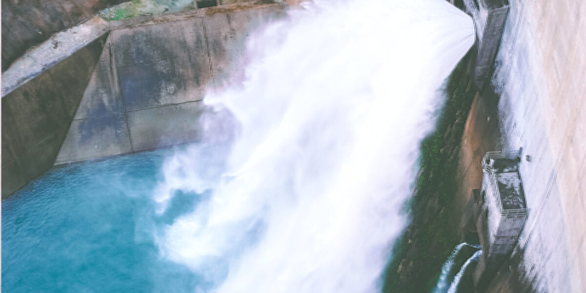 Water cascades forcefully through an open dam gate into a blue reservoir below, creating mist and turbulence. Concrete walls and structures frame the scene from above.
