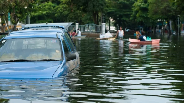 Une voiture bleue est partiellement submergée par les eaux dans une rue de la ville, tandis que des personnes à bord de petits bateaux naviguent sur l'eau à l'arrière-plan, entourées d'arbres et de bâtiments.