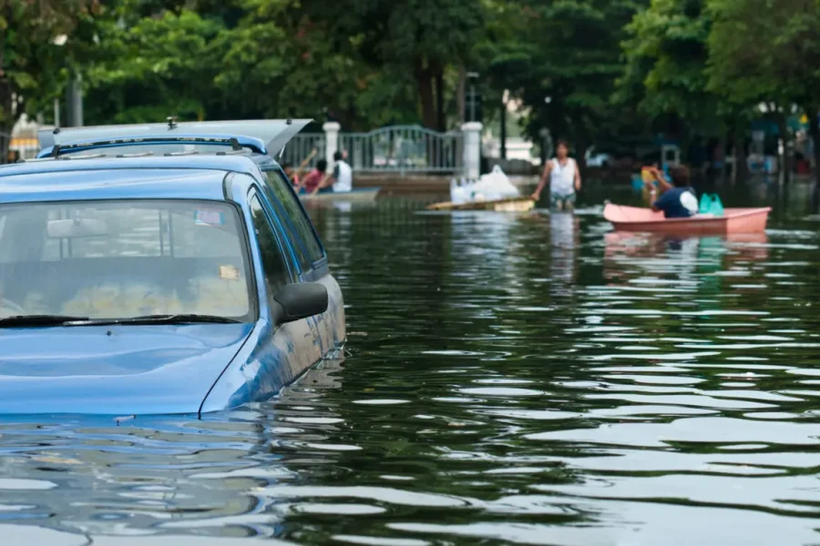 Une voiture bleue est partiellement submergée par les eaux dans une rue de la ville, tandis que des personnes à bord de petits bateaux naviguent sur l'eau à l'arrière-plan, entourées d'arbres et de bâtiments.