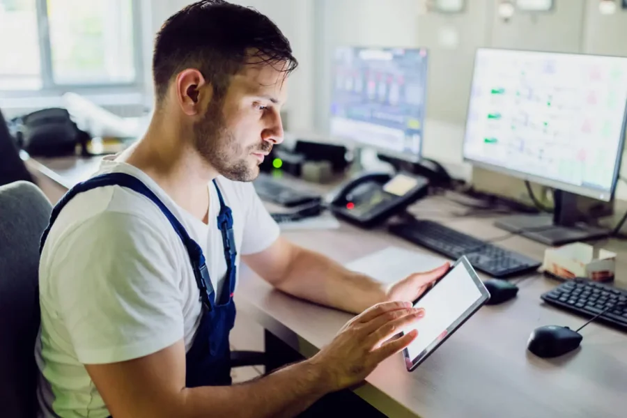 Un homme en bleu de travail est assis à un bureau avec plusieurs écrans et équipements informatiques, utilisant une tablette dans une salle de contrôle ou un environnement de bureau.