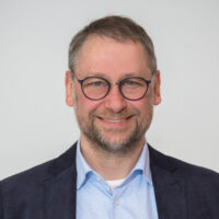 A middle-aged man with short brown hair, a beard, and glasses is smiling. He is wearing a light blue collared shirt, a white undershirt, and a dark jacket featuring the HyQuant logo. The background is plain and light-colored.