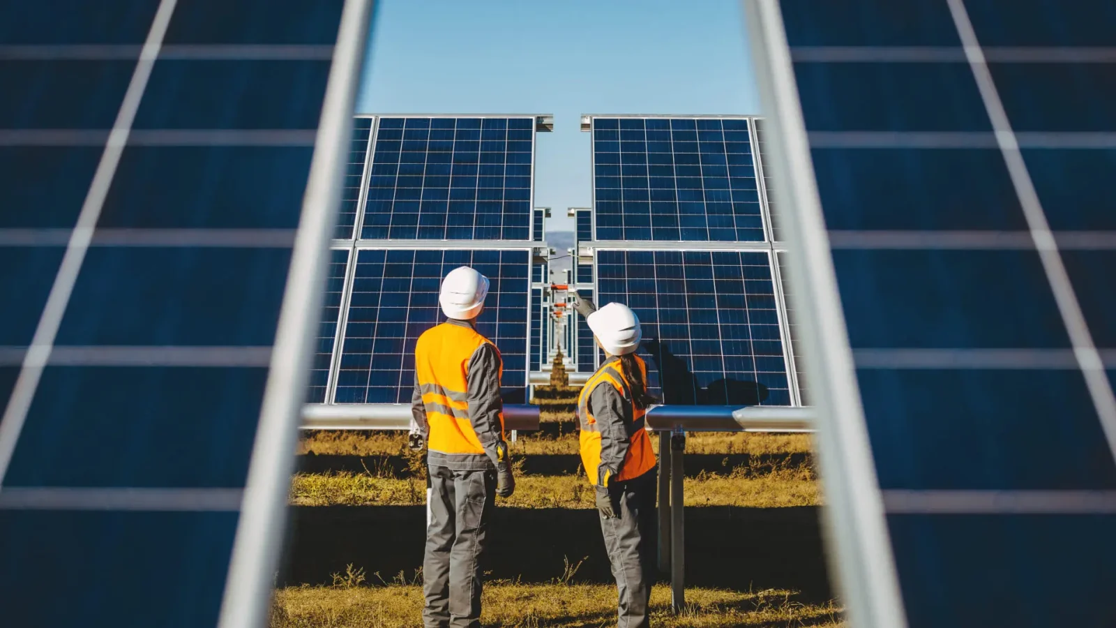 Deux ouvriers vêtus de gilets de sécurité et de casques orange se tiennent face à de grands panneaux solaires en plein air, d'autres panneaux étant visibles au premier plan sous un ciel bleu clair.