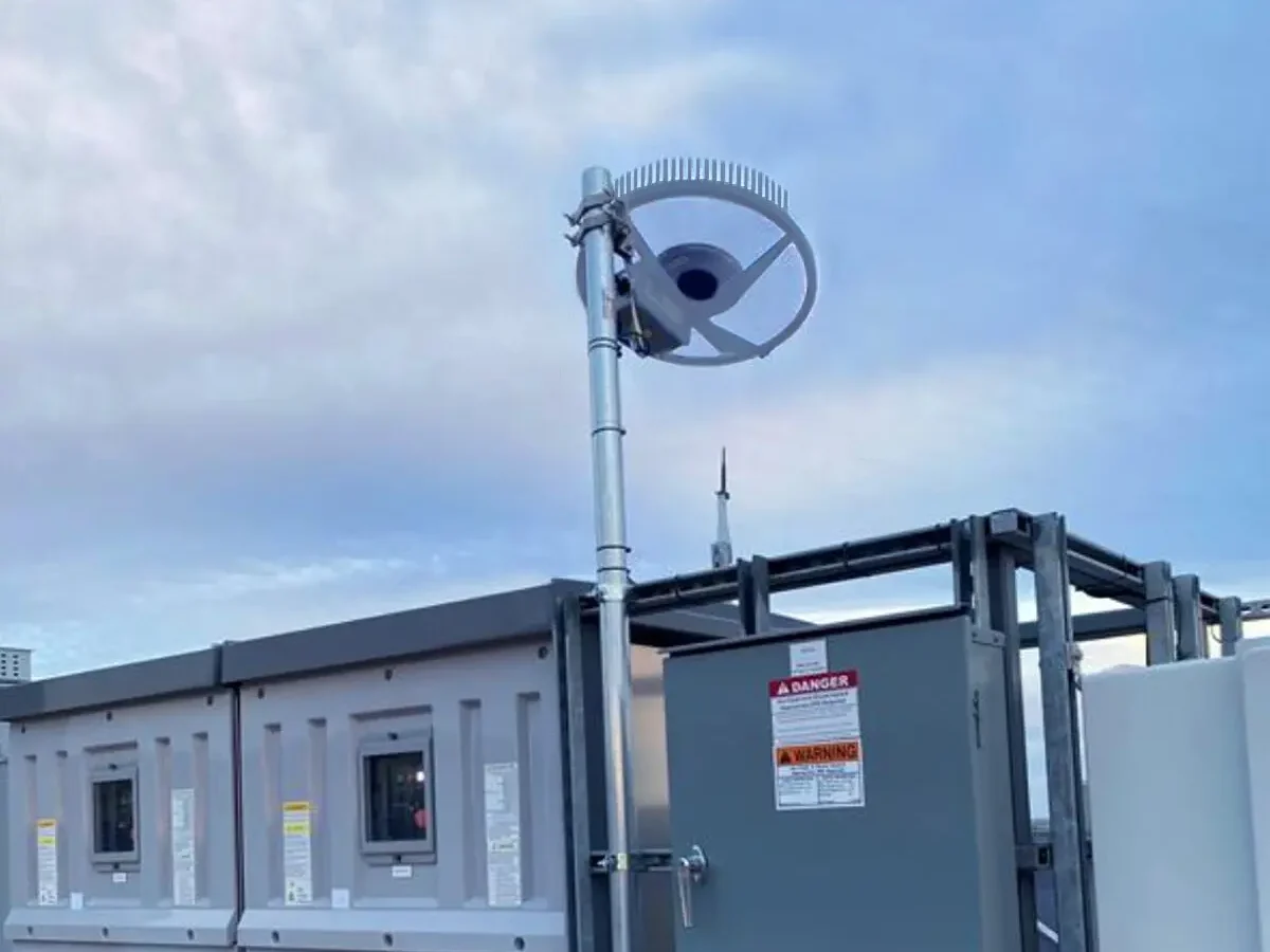 Une antenne parabolique circulaire Starlink est montée sur un poteau métallique à côté d'une boîte de service public grise avec des panneaux d'avertissement, sur fond de ciel nuageux.