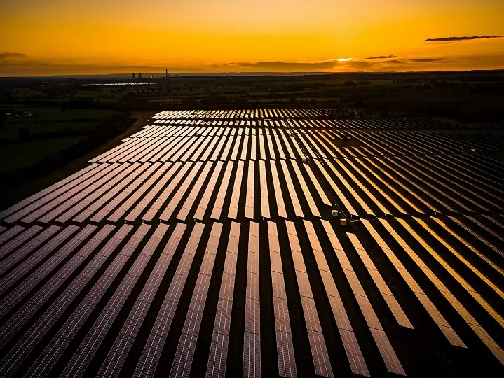 Une grande ferme solaire avec des rangées de panneaux solaires reflétant la lumière orange du coucher de soleil, dans un paysage rural avec des champs et des arbres lointains.