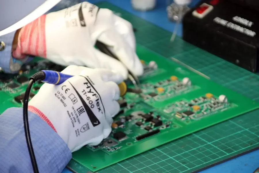 Une personne portant des gants blancs soude des composants sur un circuit imprimé vert à l'aide d'un fer à souder sur un établi recouvert d'un tapis de coupe.