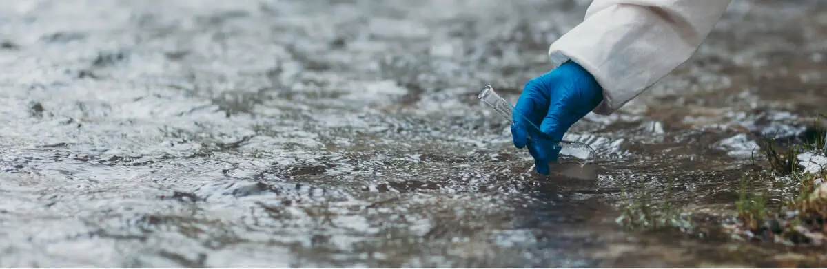 Une personne portant une blouse blanche et un gant bleu prélève un échantillon d'eau dans un ruisseau ou une rivière à l'aide d'une petite fiole en verre.
