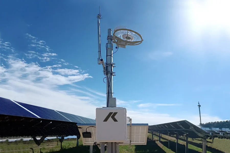 Une station météorologique équipée de capteurs et d'antennes se trouve à côté de rangées de panneaux solaires sous un ciel bleu ensoleillé avec des nuages épars. L'herbe verte entoure le réseau de panneaux solaires.