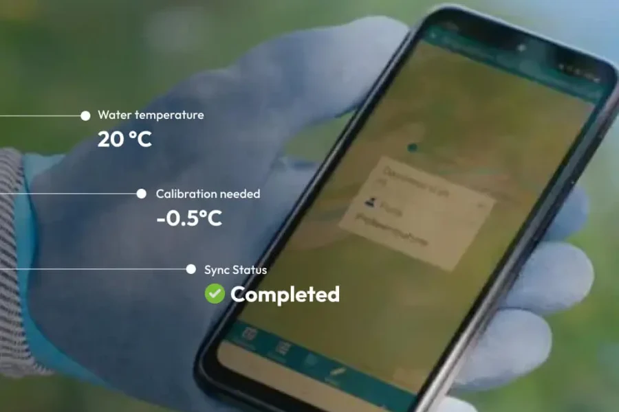 Une main gantée tient un smartphone affichant les données de surveillance de l'eau : température de l'eau à 20 °C, étalonnage nécessaire à -0,5 °C et état de la synchronisation marqué d'une coche verte.