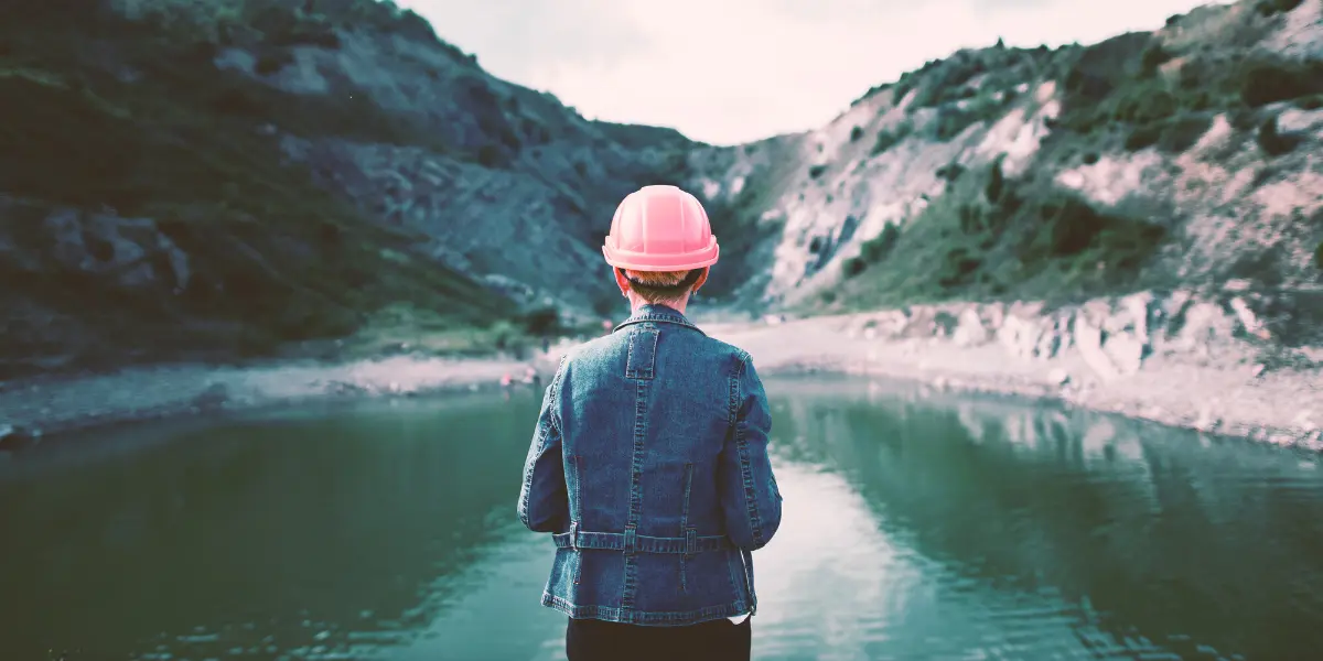 Une personne portant un casque rose et une veste en jean se tient au bord d'un lac calme, entouré de collines rocheuses et de verdure, face à la caméra.