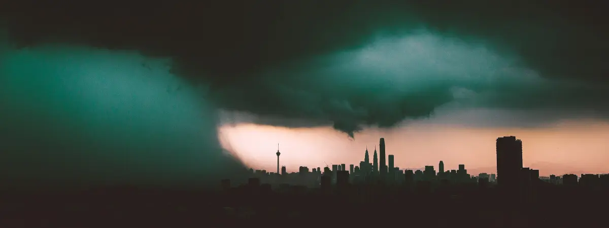 De sombres nuages d'orage se profilent au-dessus de la ligne d'horizon d'une ville au crépuscule, la pluie tombant d'un côté et plusieurs grands bâtiments se détachant sur une faible lueur orange dans le ciel.