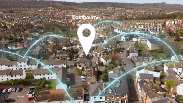 Vue aérienne d'un quartier résidentiel d'Eastbourne avec une épingle de localisation numérique et des lignes bleues soulignant la connectivité entre les maisons et les rues, sur fond de collines.