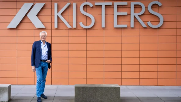 Un homme vêtu d'un blazer bleu et d'un jean se tient, souriant, devant un mur orange moderne surmonté de l'enseigne KISTERS et d'un logo stylisé. Deux bancs en béton se trouvent devant le mur.