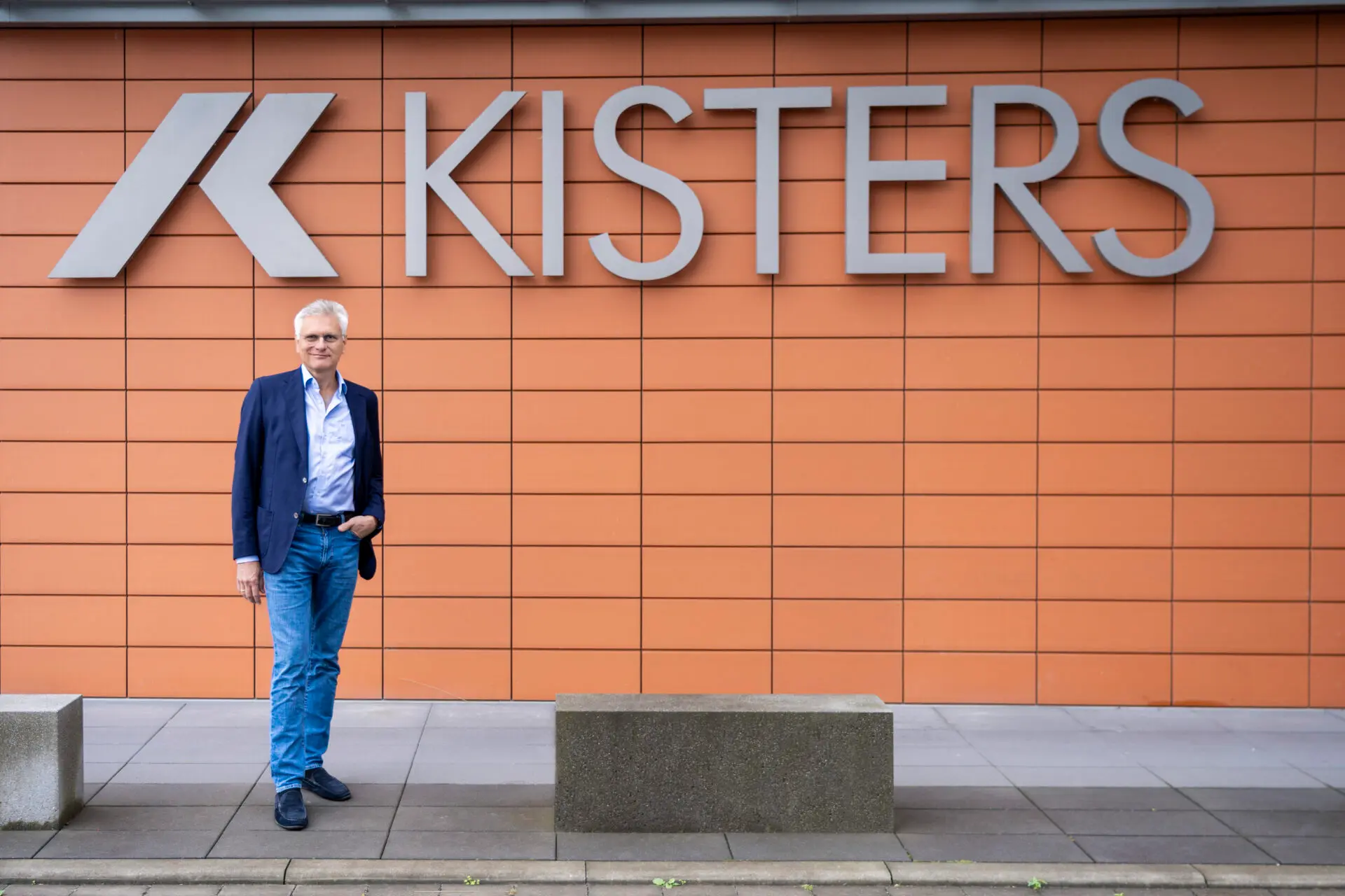 A man in a blue blazer and jeans stands in front of an orange tiled wall with a large KISTERS sign and logo. Concrete benches are on the pavement beside him.