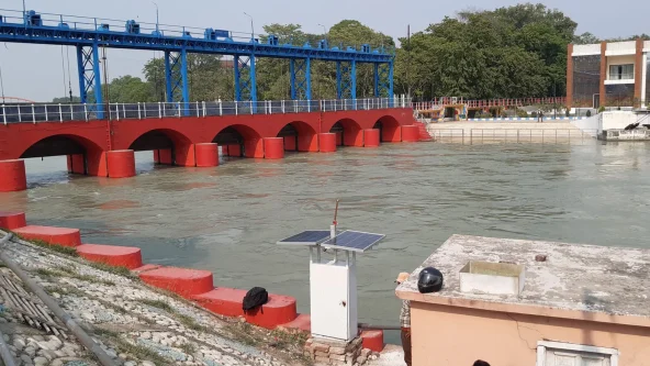 Un barrage hydraulique avec des arches rouges et des grilles bleues enjambe une rivière. L'eau s'écoule à travers les arches. Un petit bâtiment avec un panneau solaire se trouve au premier plan et des arbres à l'arrière-plan.