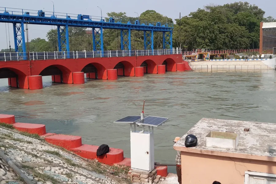Un barrage hydraulique avec des arches rouges et des grilles bleues enjambe une rivière. L'eau s'écoule à travers les arches. Un petit bâtiment avec un panneau solaire se trouve au premier plan et des arbres à l'arrière-plan.