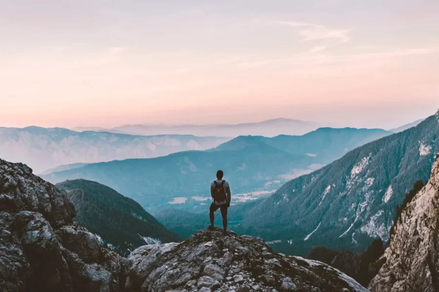 Une personne munie d'un sac à dos se tient sur un affleurement rocheux, surplombant un vaste paysage de montagnes et de vallées sous un ciel aux couleurs pastel au lever ou au coucher du soleil.