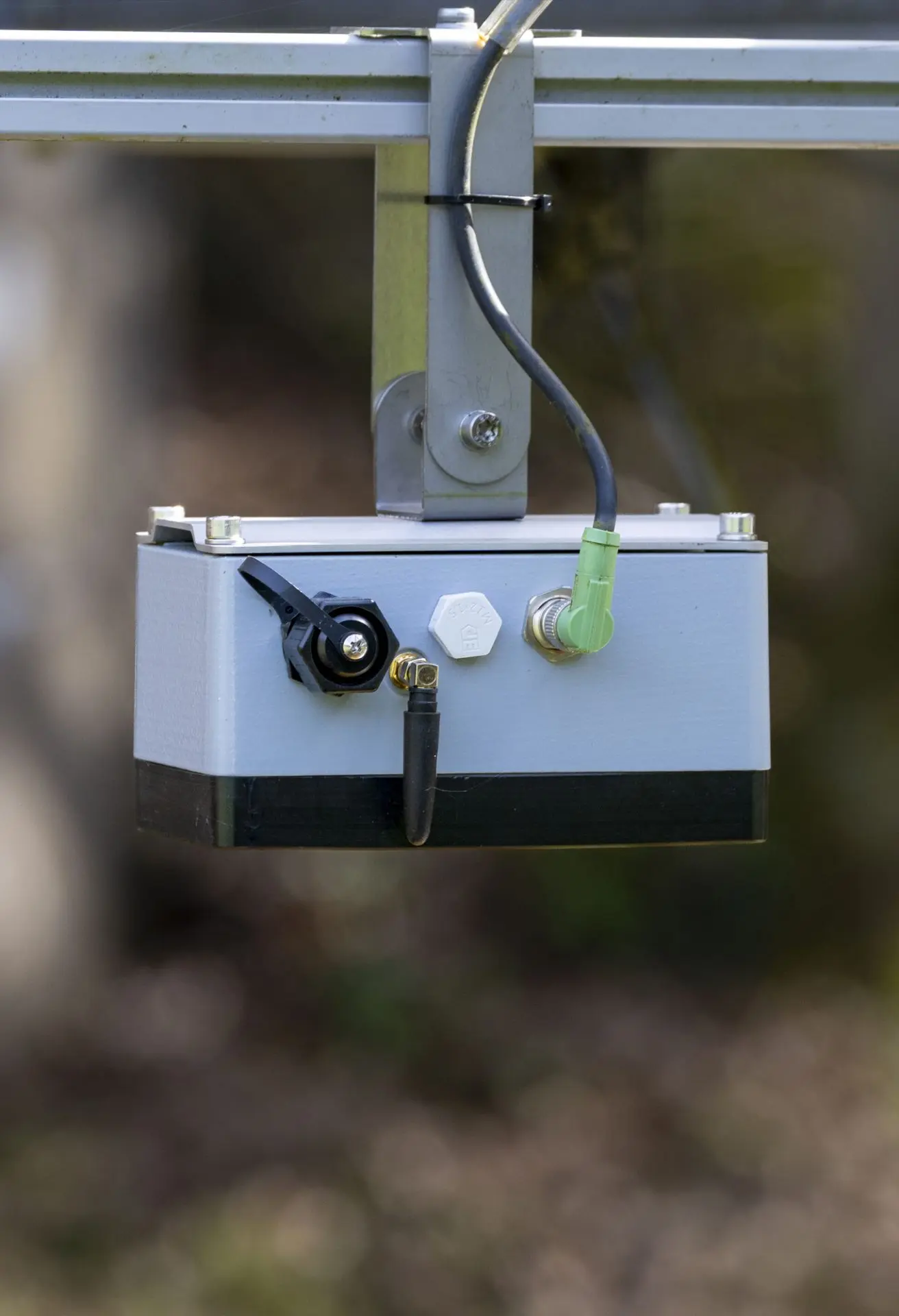 Gros plan d'un boîtier de capteur électronique d'extérieur monté sur un cadre métallique, avec plusieurs fils et connecteurs attachés, sur un fond naturel flou.