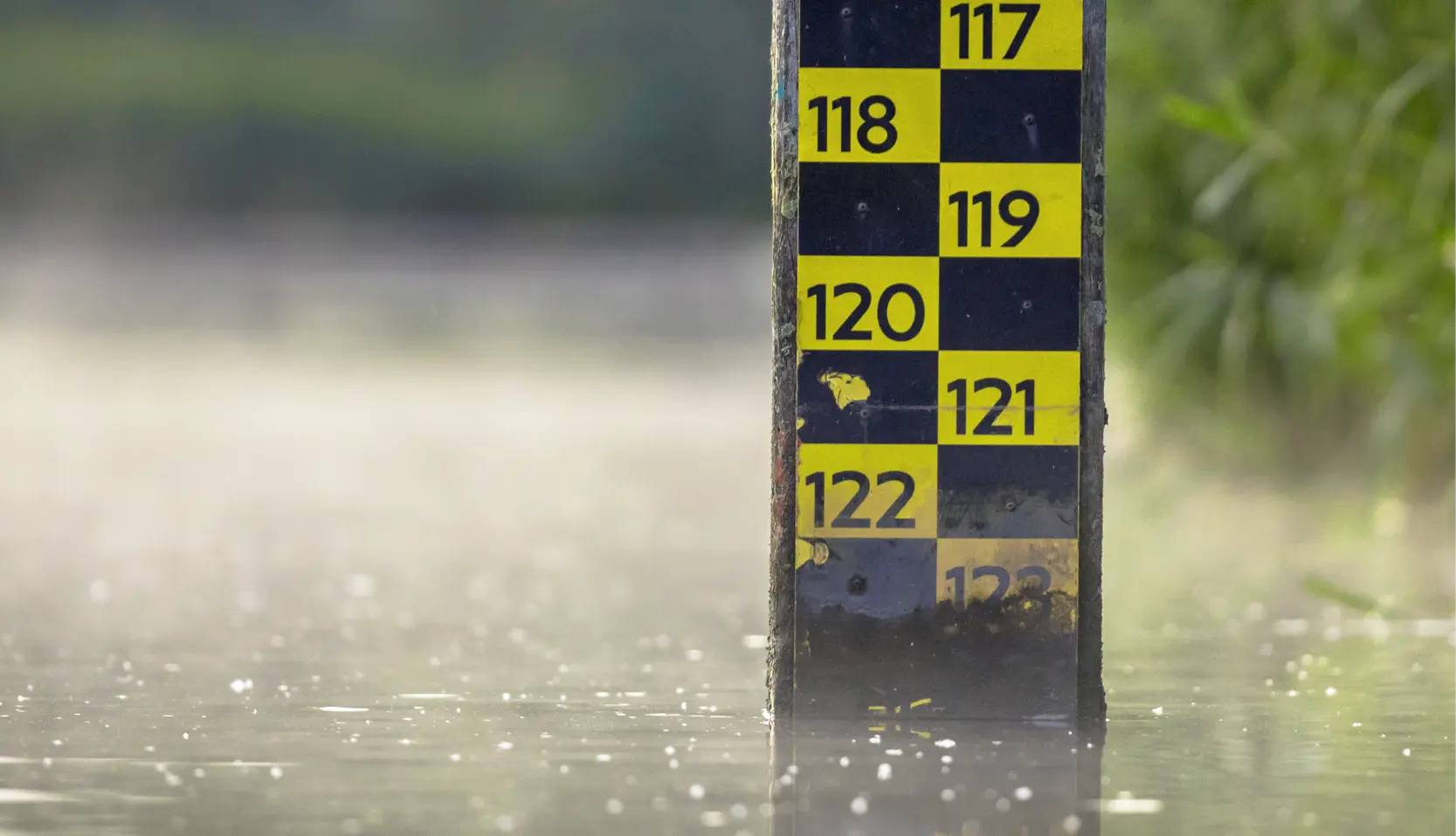 Gros plan d'une jauge de niveau d'eau marquée de chiffres jaunes et noirs allant de 117 à 122, partiellement immergée dans l'eau, sur un fond naturel flou.